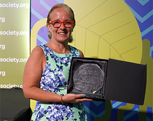 Mary Ann Torres holds her 2026 Elizabeth Taylor Human Rights Award, smiling in red glasses and a floral dress