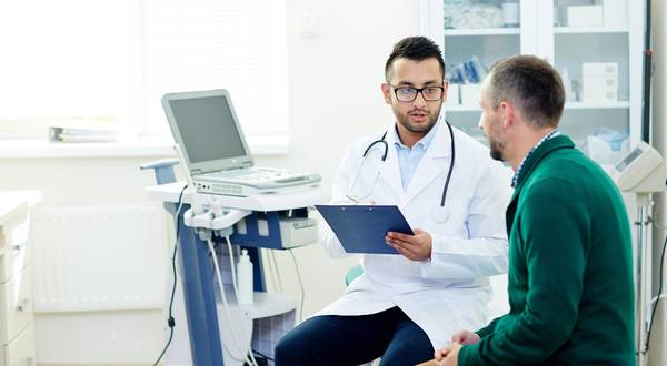A doctor in a white coat and stethoscope holds a clipboard and talks with a male patient in a bright medical exam room.