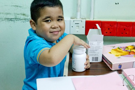 Young boy holding a medicine bottle at a desk with paperwork, medication box, and colorful folder visible nearby